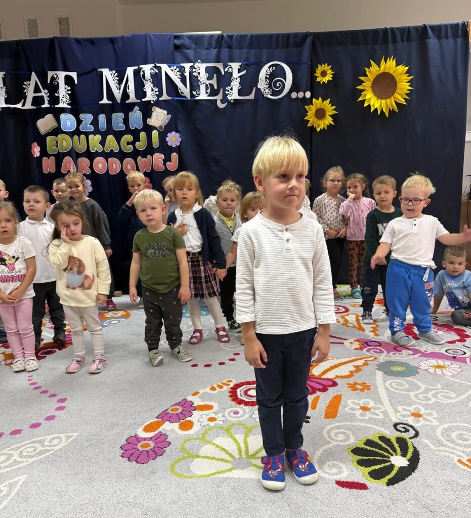 Chłopiec w jasnej koszuli stoi przed grupą dzieci na tle dekoracji Dzień Edukacji Narodowej.