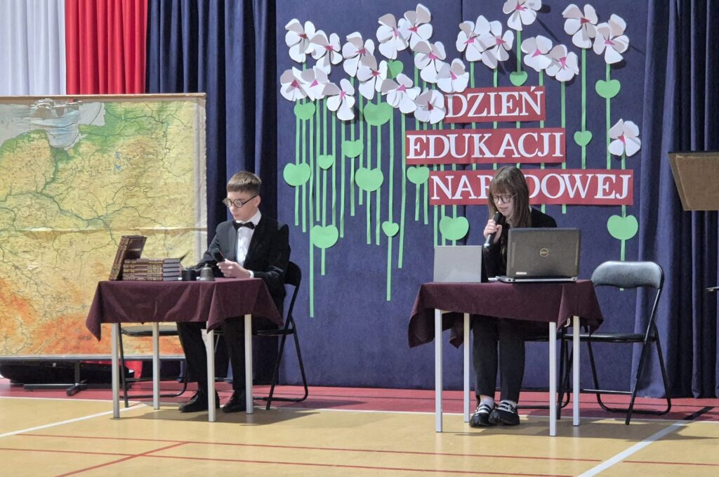 Uczniowie przy stolikach odgrywają scenkę na tle dekoracji Dzień Edukacji Narodowej.