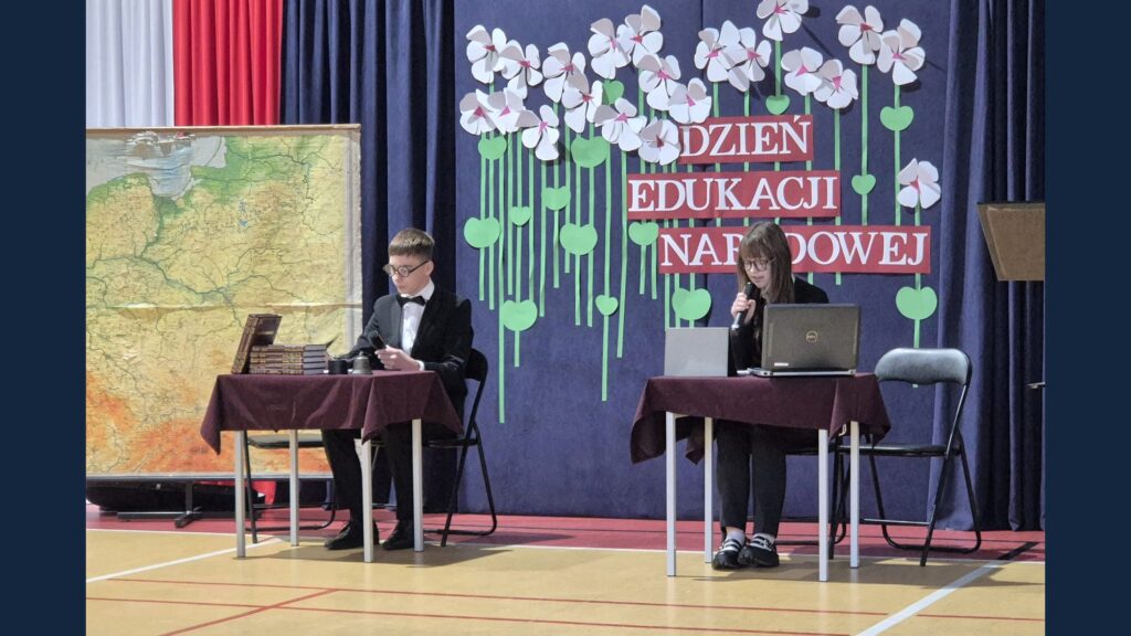 Uczniowie przy stolikach odgrywają scenkę na tle dekoracji Dzień Edukacji Narodowej.